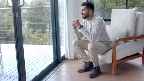 Happy-biracial-man-sitting-in-armchair-and-looking-through-window-at-home,-slow-motion