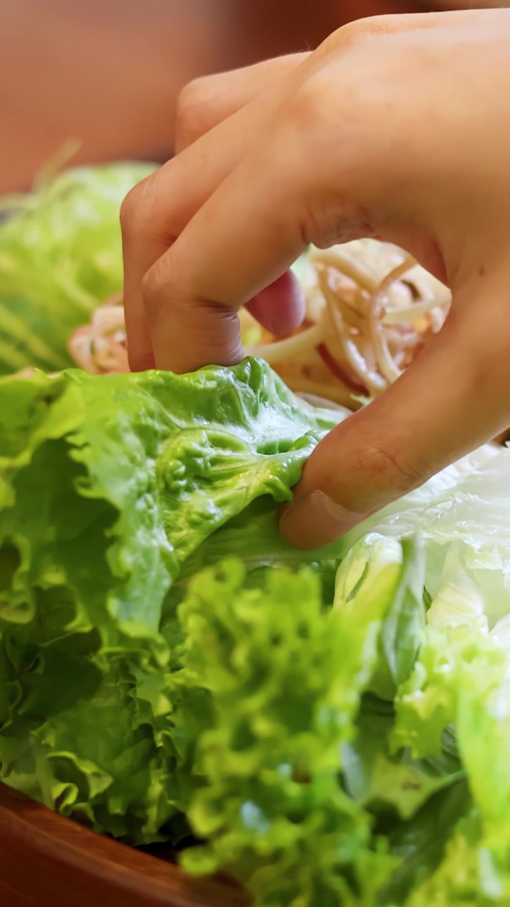 A close-up shot shows hands assembling fresh herbs and grilled pork sausage on lettuce leaves