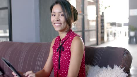 Portrait-of-happy-biracial-casual-businesswoman-using-tablet-on-couch-in-office-lounge,-slow-motion