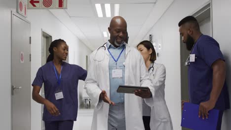 video of four diverse male and female doctors discussing tablet and files, walking in corridor