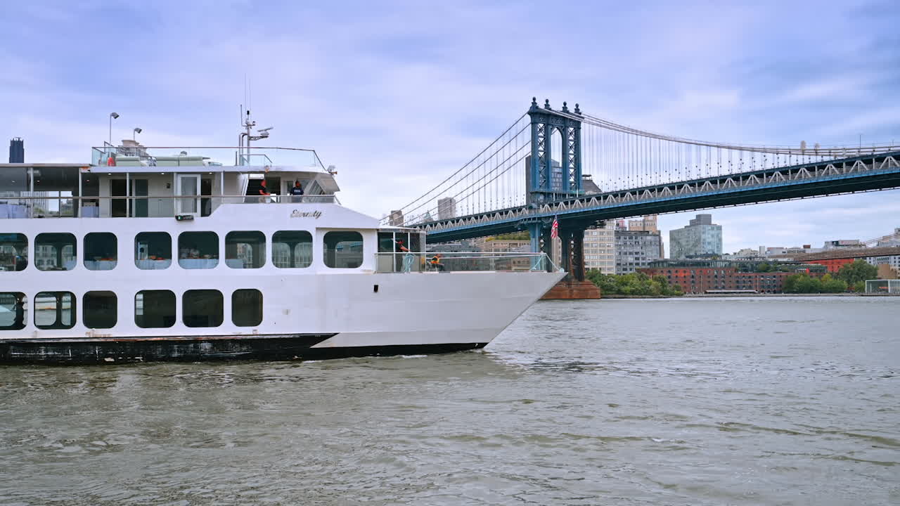 White multi-deck boat slowly moves by the riverscape. Vehicle approaches the Manhattan Bridge on a grey day
