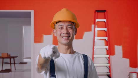 close up de hombre asiático pintor con casco de seguridad sonriendo y tocando su pecho luego apuntando a usted mientras está de pie con la pared de pintura en la habitación