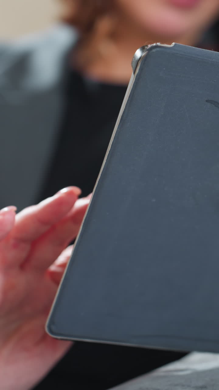 Businesswoman in grey suit holding digital tablet while gesturing with other hand, possibly during video call or touchscreen interaction, sitting indoors in calm environment with focus on productivity