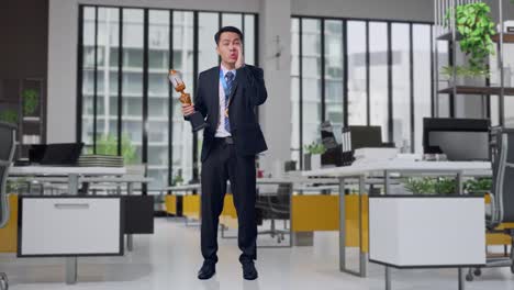 Full Body Of Asian Business Man In A Suit And Tie With A Gold Medal Holding A Gold Trophy And Yelling Positively With Hand Over Mouth In Office
