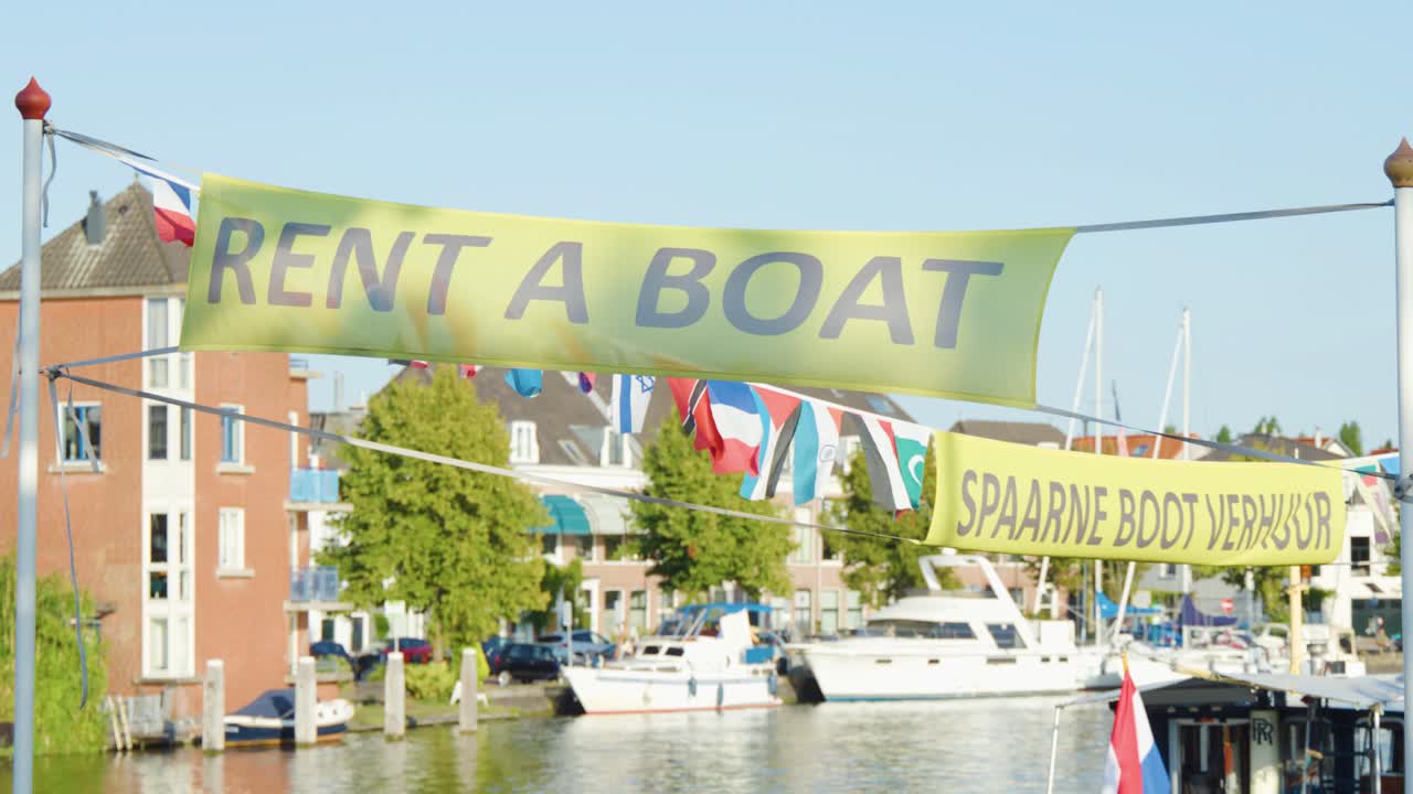 Daytime canal scene in Haarlem, Netherlands with 'Rent a Boat' banner, boats, and flags