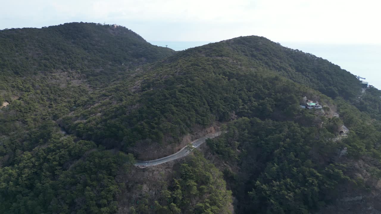 Drone aerial view in South Korea countryside flying over the dark blue sea of Busan towards green mountain with roads on a sunny day