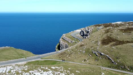 Excursionistas-Turísticos-A-Pie-En-La-Pintoresca-Carretera-Rural-De-Montaña-Verde-Con-Vistas-A-La-Hermosa-órbita-Aérea-Azul-Del-Mar-Irlandés-A-La-Derecha