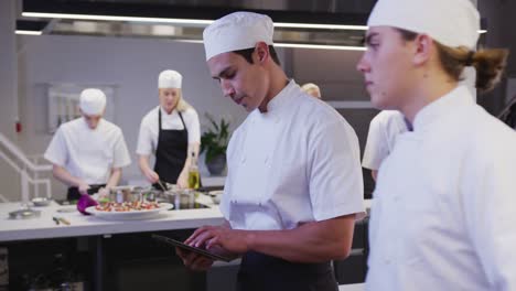 Mixed-race-male-chef-wearing-chefs-whites-in-a-restaurant-kitchen-using-a-tablet-and-looking-at-came