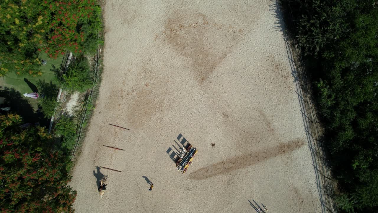 Slow aerial descent uncovers a training track where several riders perform jumps under coach observation