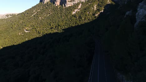 Ascending-aerial-view-of-scenic-mountainside-road-under-shadows-surrounded-by-lush-pine-trees-and-tall-mountains