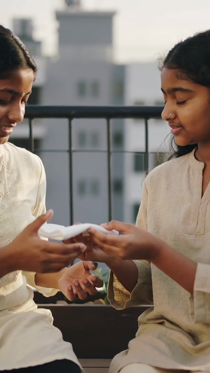 Vertical video: Lifting white cloth, right girl extending to left girl on roof bench