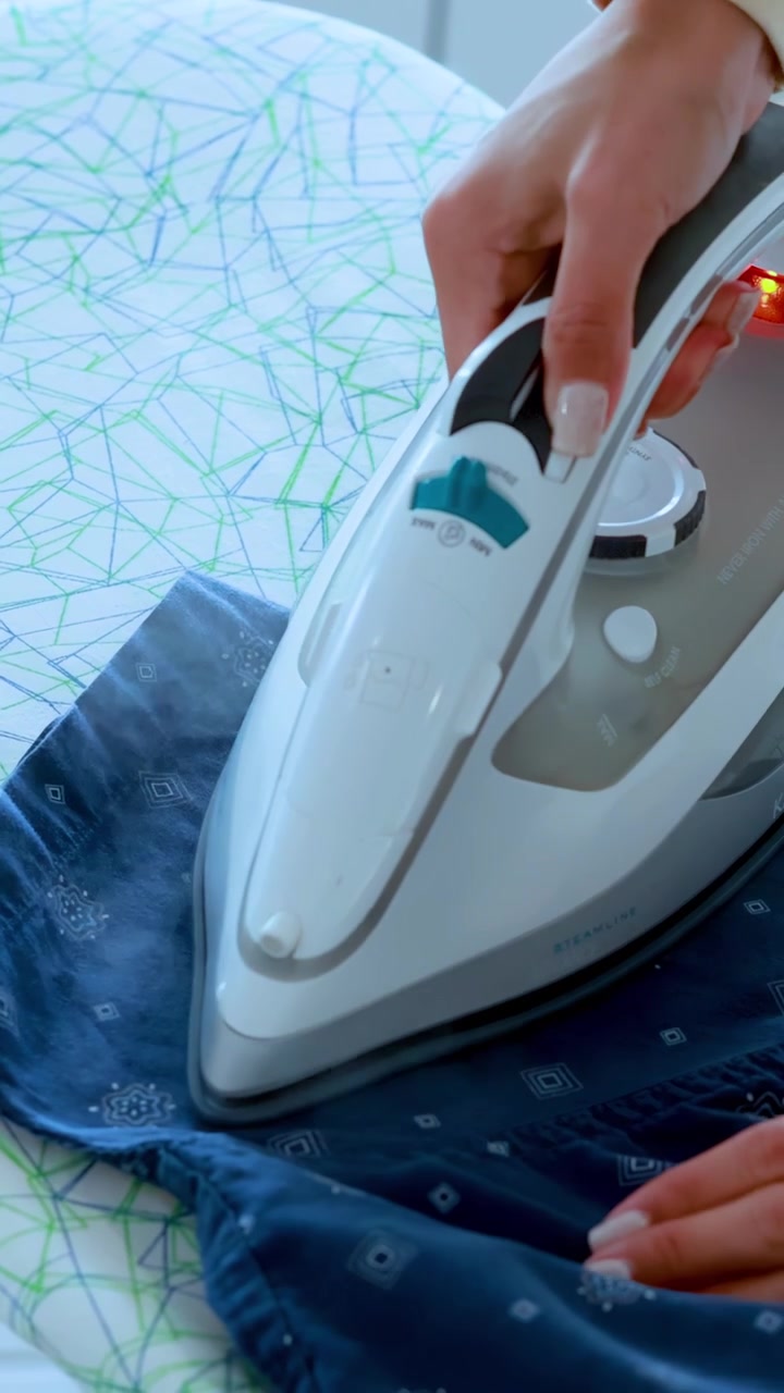 A woman uses a steam iron to press a blue patterned shirt on a modern ironing board in bright, natural lighting with close-up camera angles