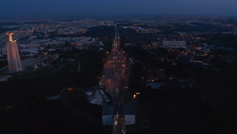 Vista-Aérea-Nocturna-De-La-Concurrida-Autopista-De-Varios-Carriles-Y-El-Monumento-Blanco-Del-Santuario-De-Cristo-Rei-En-La-Costa-De-Lisboa,-Portugal