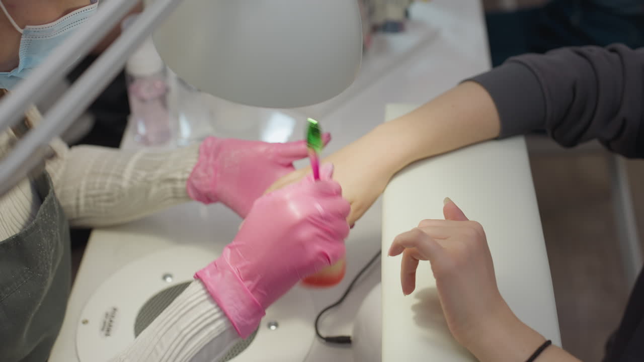 Closeup of nail technician wearing pink gloves gently brushing customer's palm and hand back using colorful soft brush under bright white lamp in clean salon setting with manicure tools visible