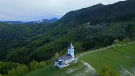 Vista-Aérea-Del-Paisaje-Montañoso-Con-La-Iglesia-Resaltada-En-Medio-De-Campos-Verdes-Y-árboles-Con-Un-Telón-De-Fondo-De-Colinas-Bajo-Un-Suave-Crepúsculo