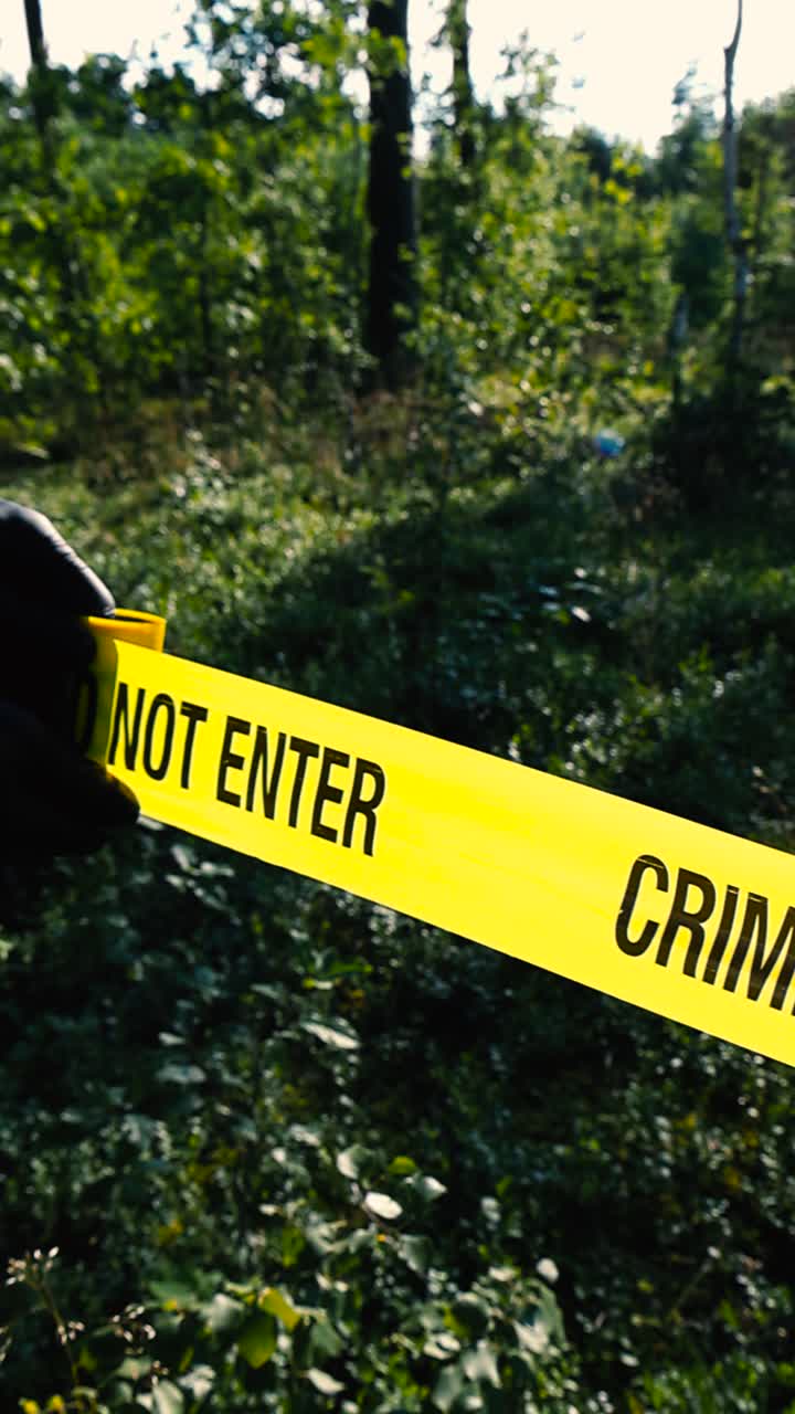 Police investigator unrolling and unraveling a forensics yellow colored Crime Scene tape in front of a wooded forest area during a summer sunny day with bokeh blurry background. Black nylon gloves