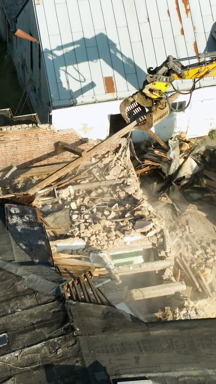 Høyt vinkelbilde av en gul gravemaskins hydrauliske klo som river ned en boligstruktur med støv og rusk. Filmatisk opptak av byfornyelse og rivningsprosess.