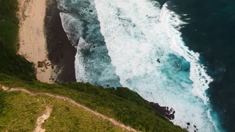 Drone-shot-of-the-famous-Kelingking-beach-with-a-side-view-of-the-steep-hiking-trail-with-waves-crashing-into-the-shoreline-on-the-island-of-Nusa-Penida,-Indonesia