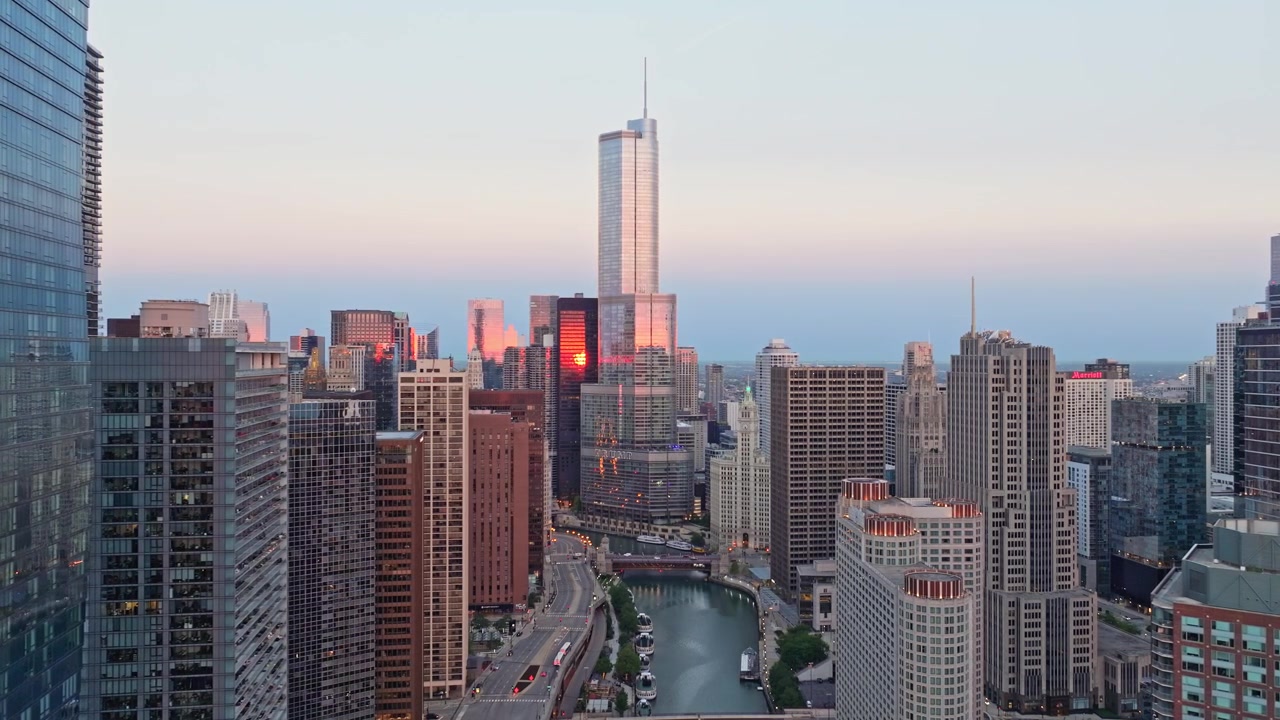 Luftfoto med drone som panorerer mot høyre over Chicago River og omkringliggende skyskrapere i sentrum som reflekterer et varmt oransje solnedgangsskjær i skumringen