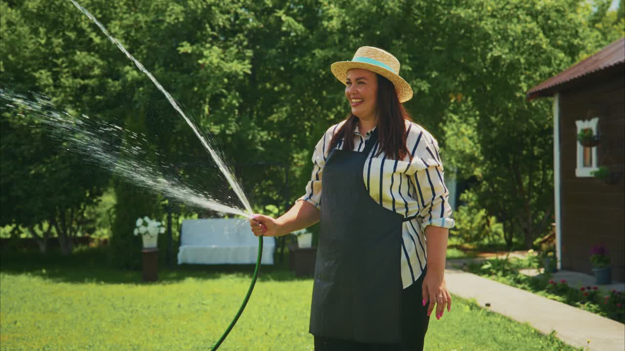 Woman watering garden with hose, wearing straw hat and apron, enjoying sunny day outdoors