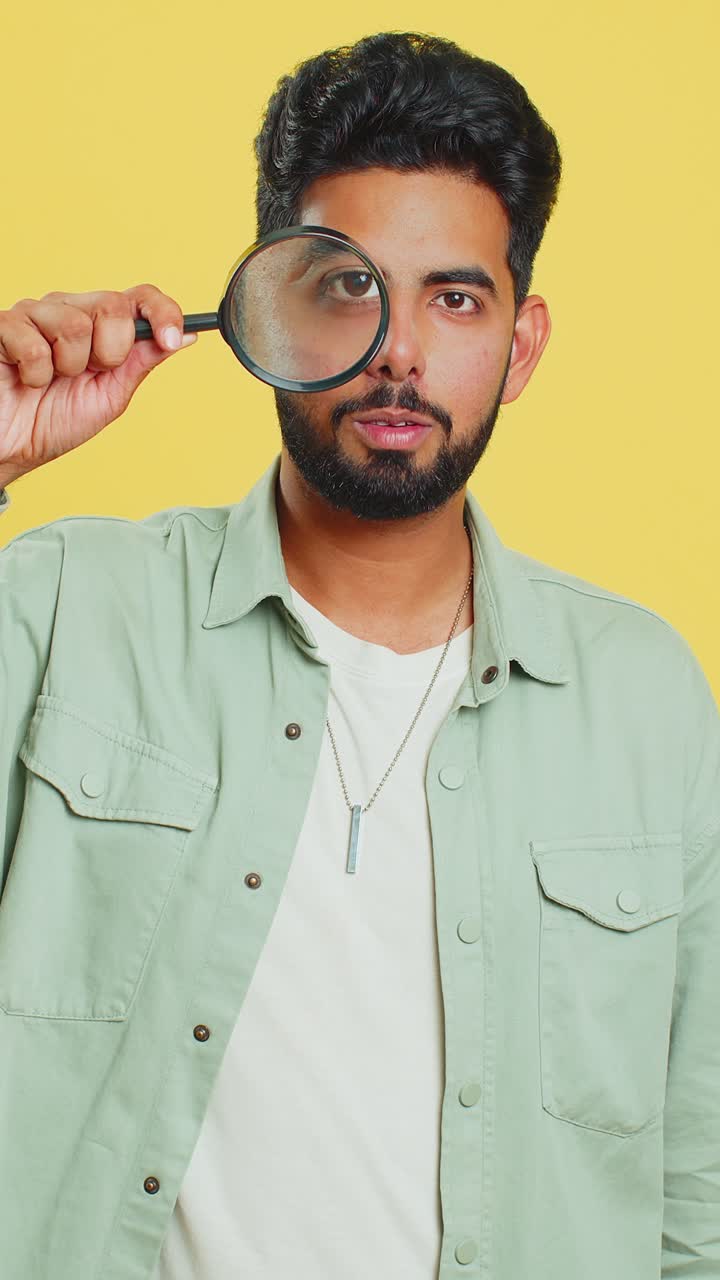 joven indio sostiene una lupa cerca de la cara mirando a la cámara con un gran ojo ampliado analizando