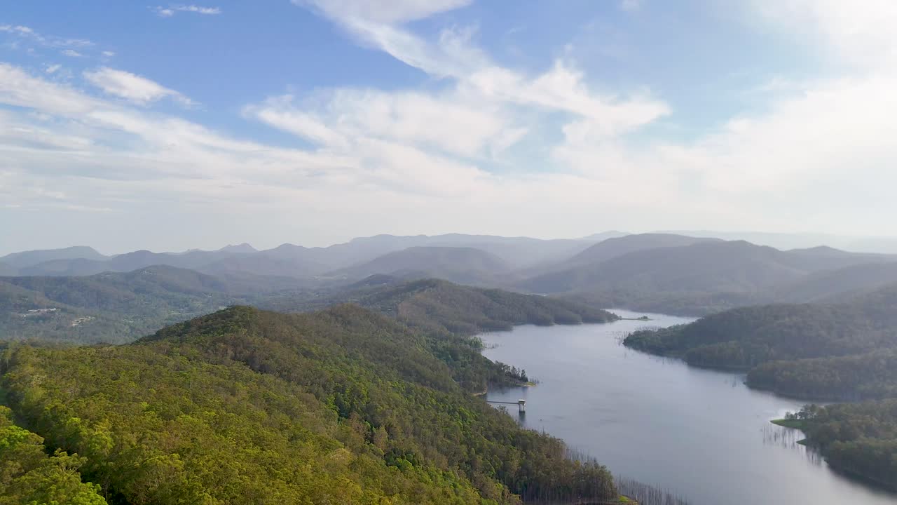Drone glides over lush forest, mountain reservoir, and distant hills in bright daylight