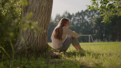 vista lateral de una mujer joven sentada en el suelo cubierto de hierba, perdida en sus pensamientos, con los brazos cruzados sobre las rodillas, la luz del sol se refleja suavemente en ella, rodeada de vegetación borrosa y tronco de árbol