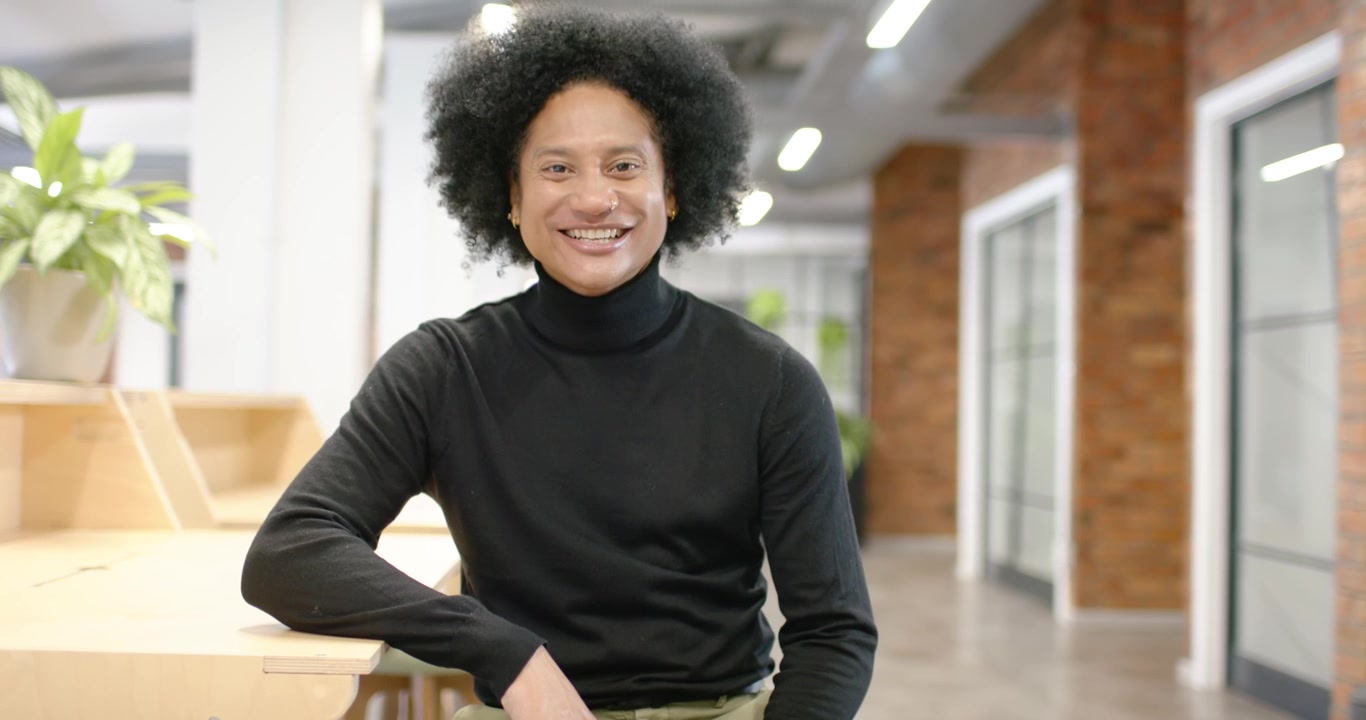 Afroamerikansk man klädd i svart sitter vid ett kontorsbord och ler mot kameran medan han spelar in en intervju.