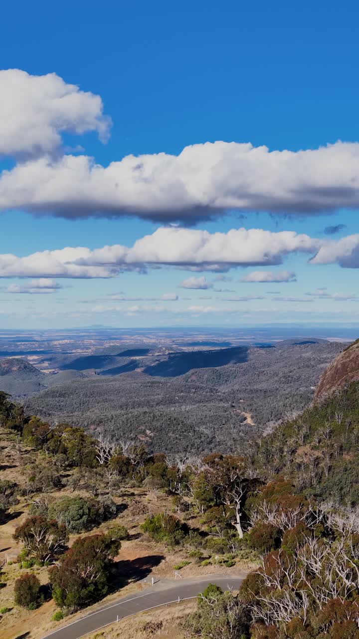 La telecamera di un drone si innalza sopra colline boscose e una strada tortuosa, rivelando ampi panorami montani sotto la piena luce del giorno e nuvole sparse nel Parco Nazionale di Warrumbungle.