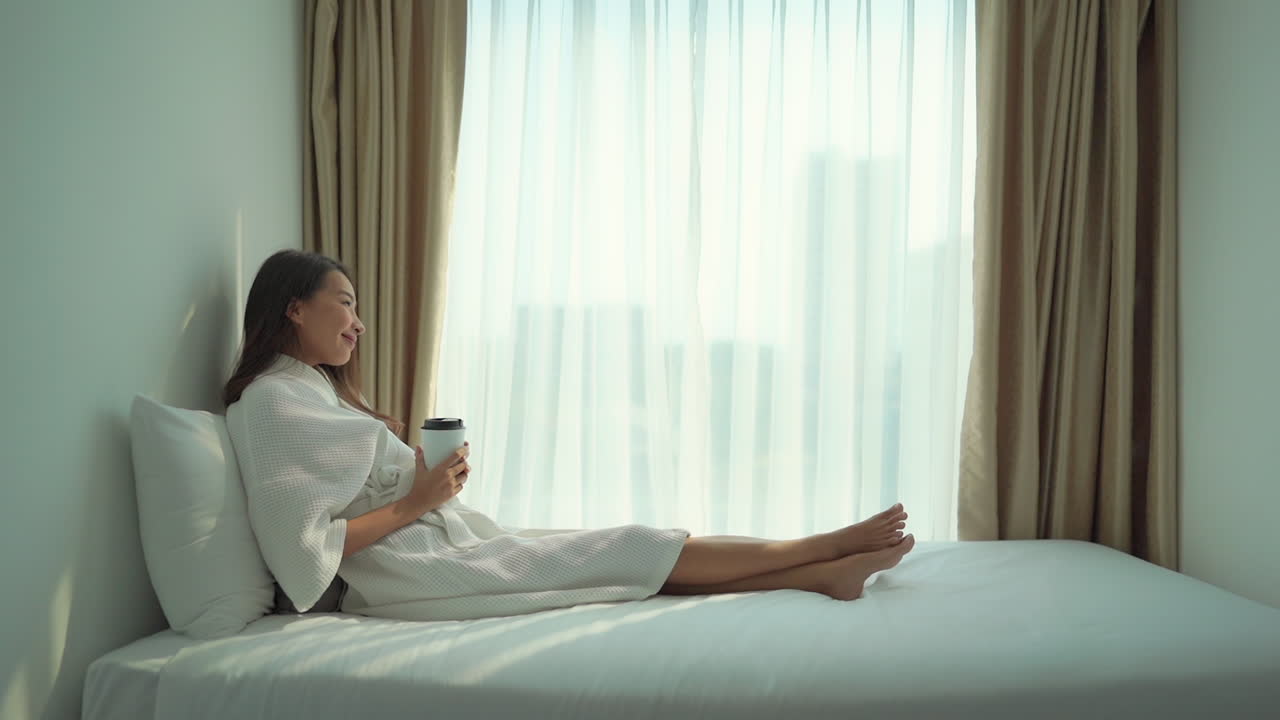 A young Asian woman dressed in a robe relaxes on the bed while preparing to drink from a disposable coffee cup