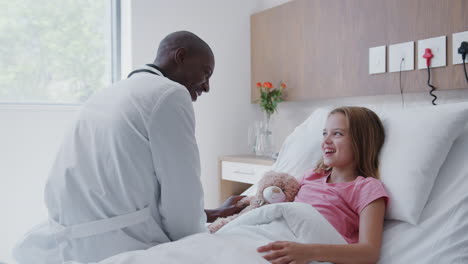 médico visitando a una chica acostada en una cama de hospital abrazando a un oso de peluche