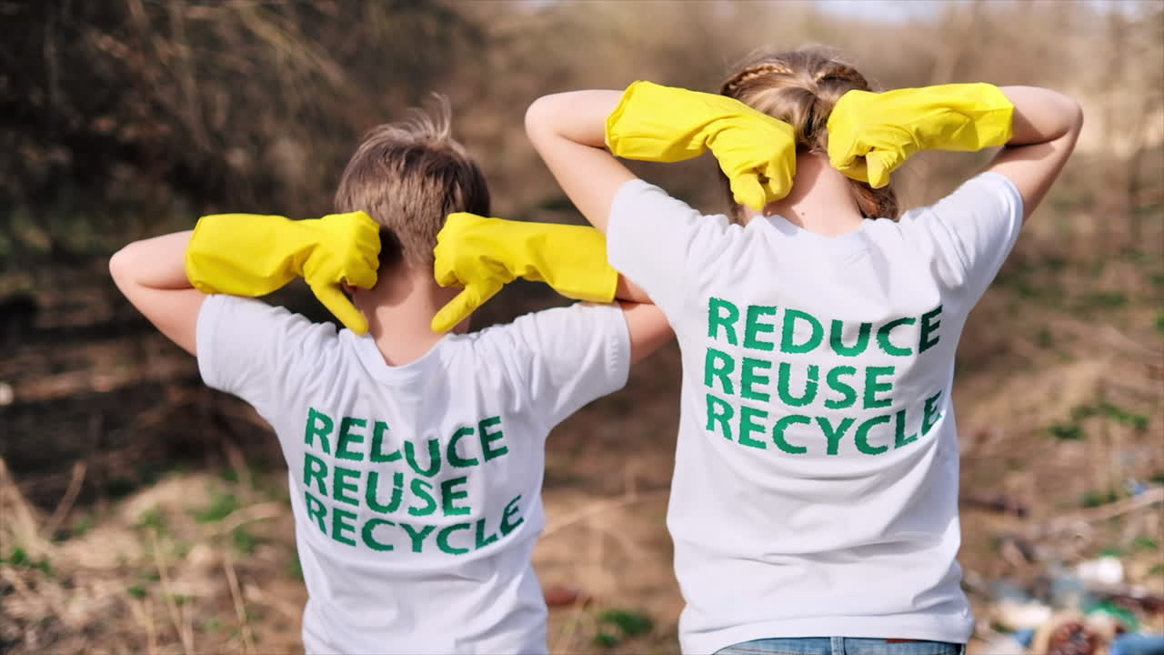 Boy and girl in rubber gloves showing on the eco inscriptions on their T-shirts. Plastic garbage collection in a polluted clearing