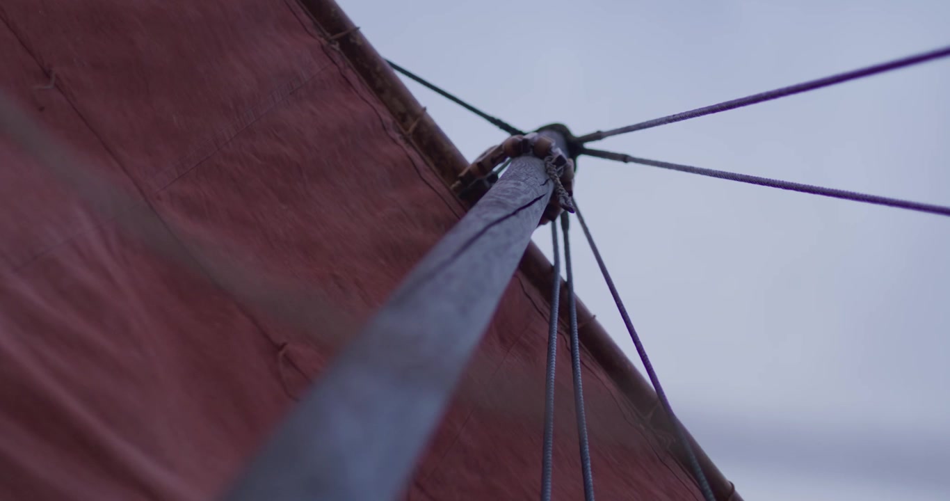 Vista dal basso di un albero di barca a vela con una vela rossa e usurata e cime scure che si estendono verso un cielo grigio-azzurro pallido, evocando avventura nautica e viaggi marittimi