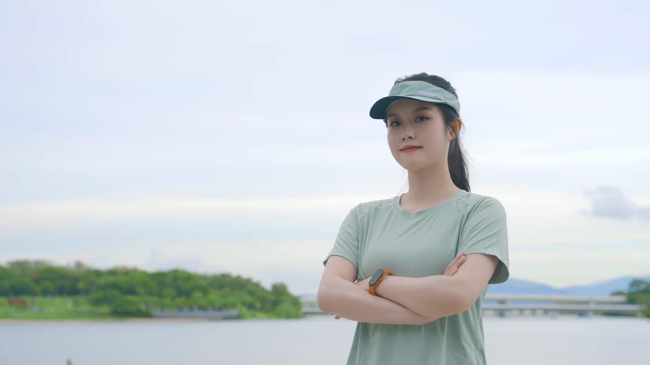 Portrait of smiling young woman with sportswear has crossed arms and fitness watch, ready for exercise. Female athlete, smile and standing at lakeside for workout, summer training or sports