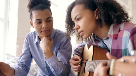 mixed race young woman with tablet computer sitting on bed teaching her teenage sister to play acoustic guitar at home