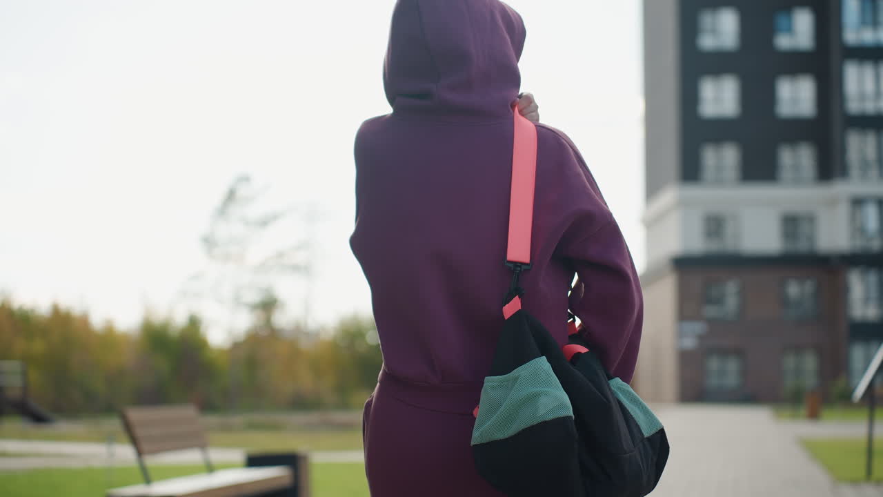 Back view of active woman walking towards building on gray tiled pavement under bright sunny sky with autumn foliage and blurred urban park surroundings capturing determined stride style