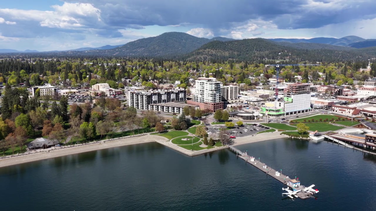 Lungolago di Coeur d'Alene con spiaggia sabbiosa, parchi verdi, moderni grattacieli residenziali, un affascinante centro città e una fitta pineta che si estende verso catene montuose blu. Ampia ripresa aerea laterale.