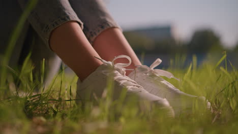 close-up of young woman s legs in jeans and white canvas shoes on lush grass, with sunlight creating a soft glow on her shoes and gentle breeze moving grass blades