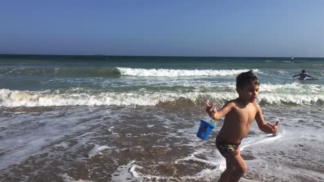 slow motion footage of caucasian toddler boy, running out of sea with big waves, holding a toy bucket in his hands