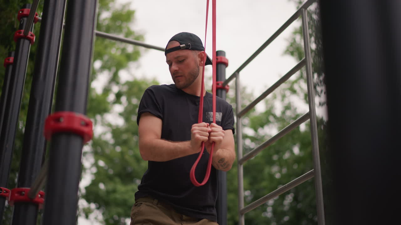 Man Securing Safety With Care, Person Carefully Tightening Safety Harness On Park Training Structure, Man Concentrates As He Adjusts Safety Rope On Outdoor Exercise Apparatus In Leafy Park