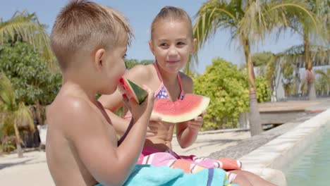 happy caucasian siblings eating watermelon at swimming pool at beach house