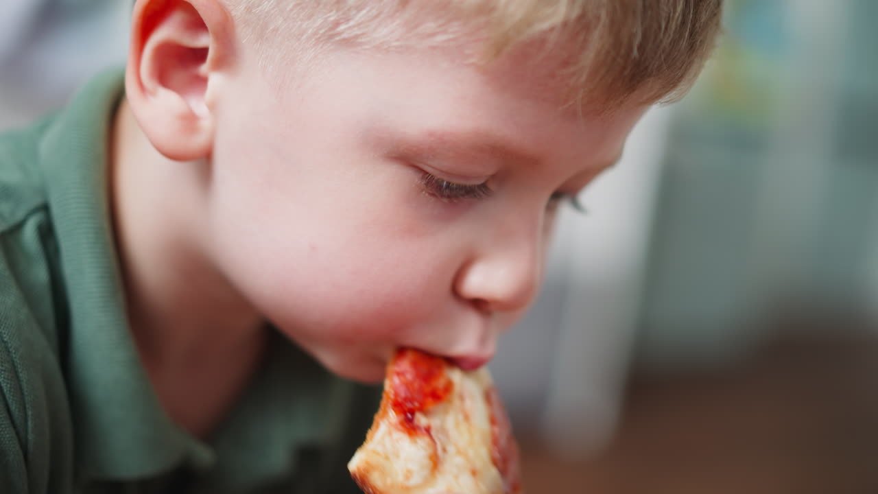 Close up of young boy in green shirt biting into cheesy pepperoni pizza with intense focus, showing joyful mealtime moment indoors with soft background