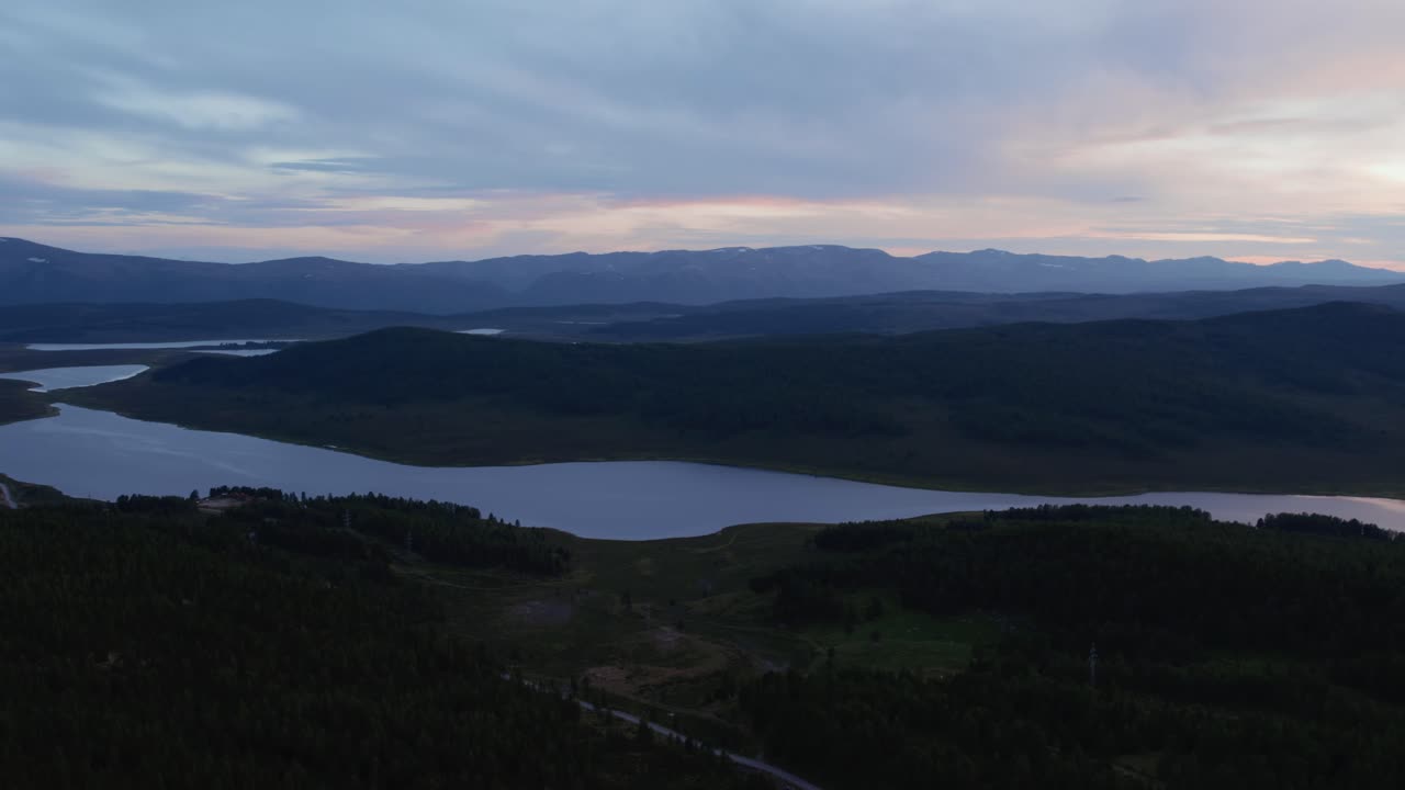 Ampie riprese aeree di un fiume tortuoso o di un bacino idrico tra scure colline boscose e catene montuose distanti al tramonto nella Repubblica dell'Altai, Russia, sotto un cielo nuvoloso