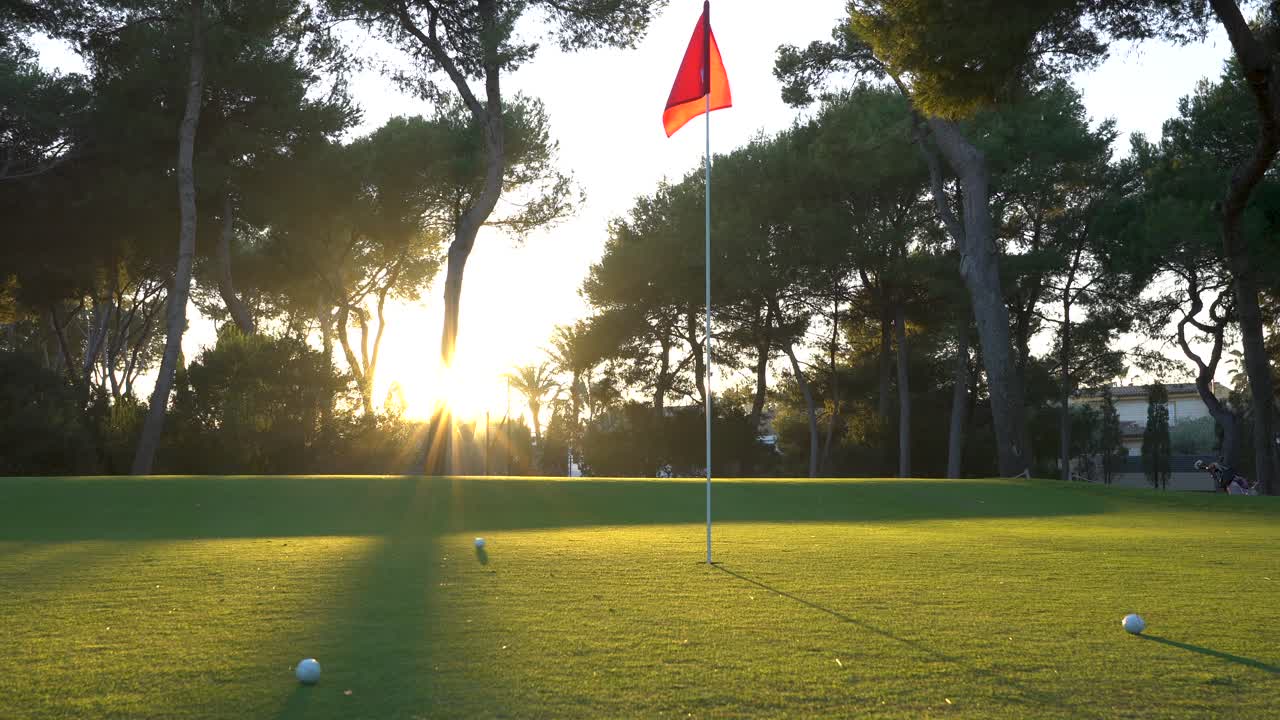 Una pallina da golf passa velocemente vicino alla bandiera, che è rossa e sventola al vento al tramonto, un bel sole illumina lo sfondo.