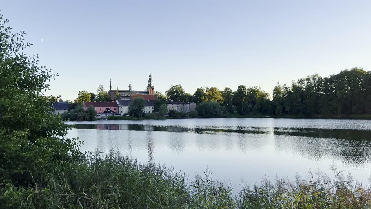 Tranquil Lakeside Scene with Gothic Church and some buildings by the lake under a soft evening sky, calm reflection on water. Historic Gothic architecture, roof and tower details are visible far away