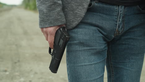 a close-up view of a man in blue jeans and a gray jacket holding a black handgun by his side while walking down a dirt road