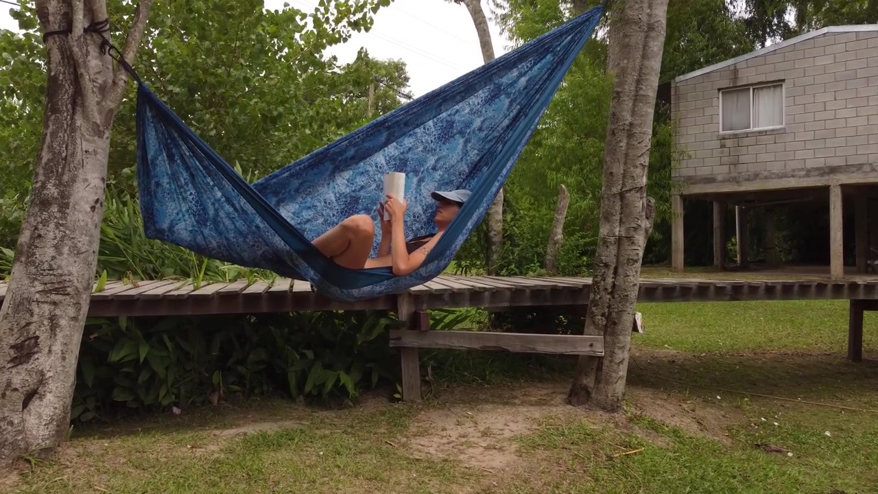 Toma dinámica de una joven leyendo un libro mientras está acostada en una hamaca azul en el patio trasero de la casa