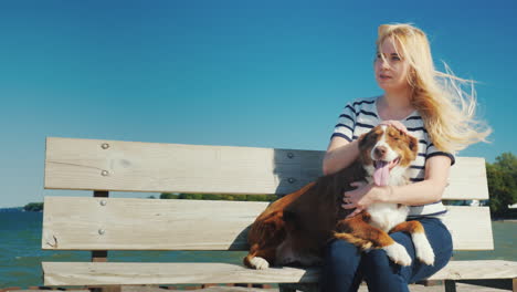 mujer joven jugando con un perro se sienta en un banco cerca del mar el viento juega con su cabello