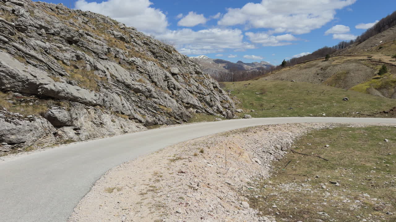 A winding, narrow mountain road snakes its way through the landscape with snow on top of the mountain under a blue sky with clouds. Sunny day, wide shot, slow motion