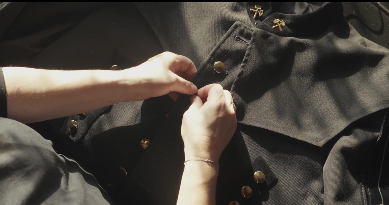 Close-up hands fasten the black tunic of a Bergmannstracht ceremonial uniform featuring crossed hammer and mallet insignia linked to mining heritage.